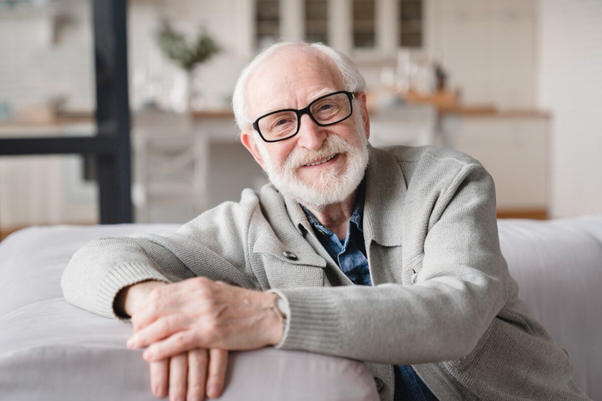 Elderly Caucasian man sitting on a couch smiling.